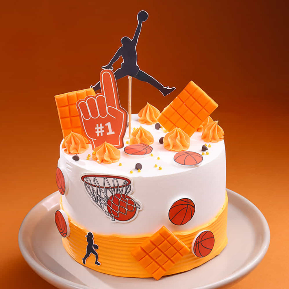 A stylish basketball-themed cake with white and orange frosting. The cake is decorated with a silhouette dunking topper, orange chocolate bars, basketball edible cutouts, and a "#1" fan foam finger, set against a warm orange background.