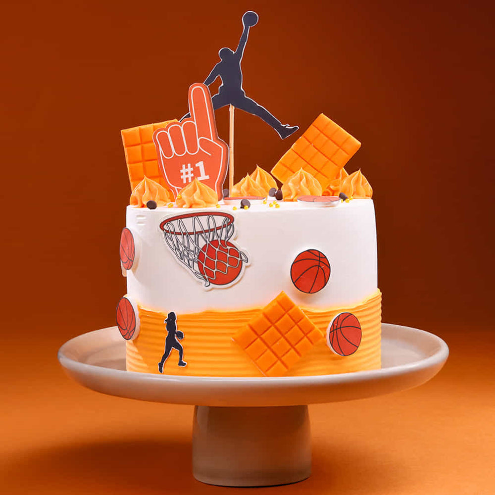 A stylish basketball-themed cake with white and orange frosting. The cake is decorated with a silhouette dunking topper, orange chocolate bars, basketball edible cutouts, and a "#1" fan foam finger, set against a warm orange background.