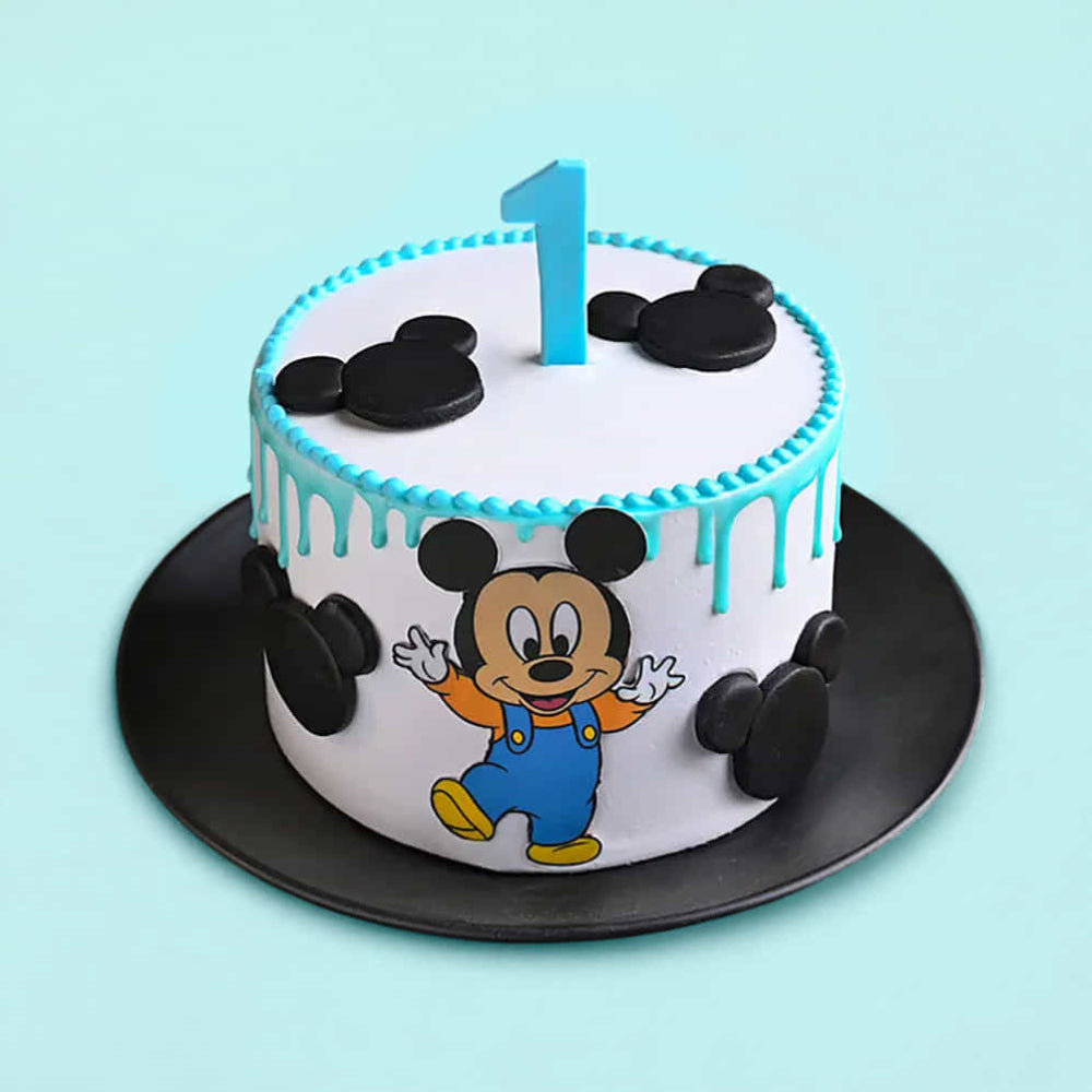 White round cake with blue drip icing, decorated with black fondant mouse head shapes and a cartoon mouse character on the side. A blue number “1” topper sits on top, and the cake is placed on a black plate against a light blue background.