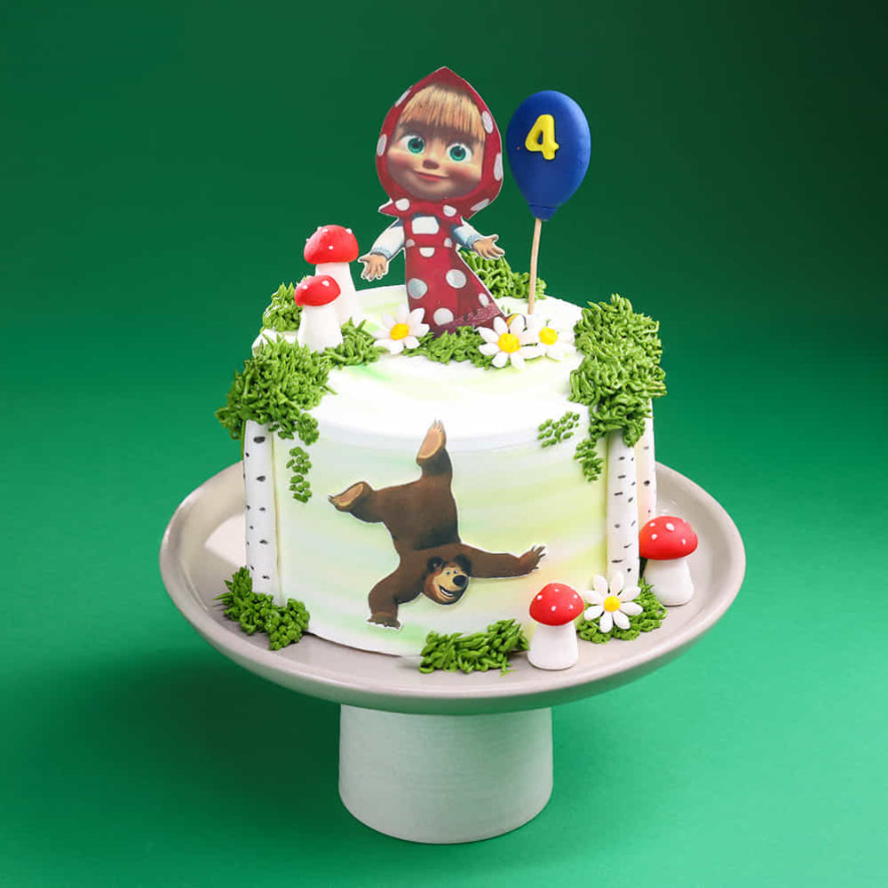 White round cake decorated with a forest theme, featuring green grass-like frosting, small red mushrooms, and flowers. On top is a cartoon girl in a red hood holding a blue balloon with the number “4,” and the side of the cake shows a playful bear illustration. The cake is displayed on a pedestal against a green background.