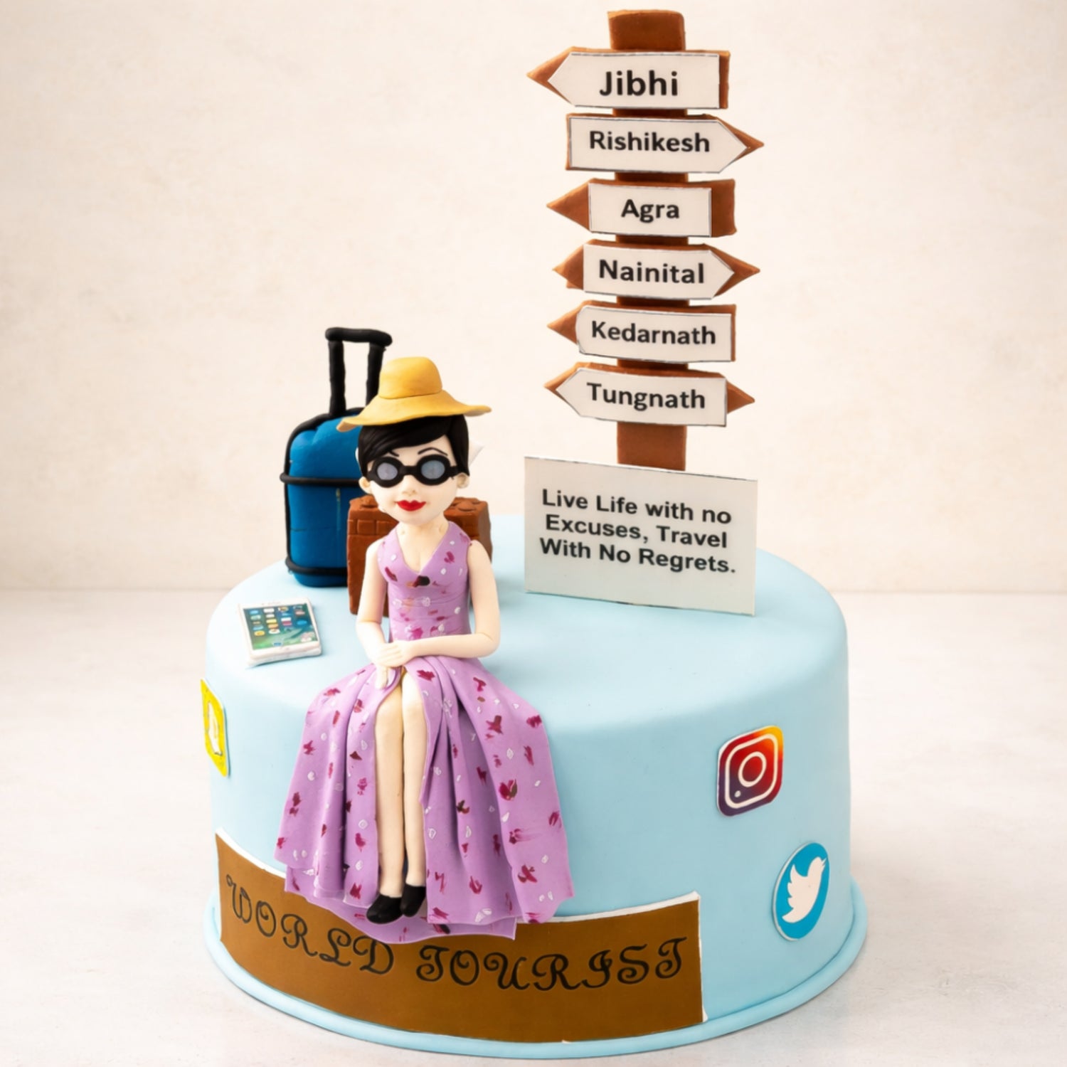 Travel-themed cake with a seated fondant figure of a woman wearing a hat and sunglasses, next to a suitcase and a signpost listing destinations. The cake features a message reading “Live Life with no Excuses, Travel With No Regrets.” and travel-related decorations.