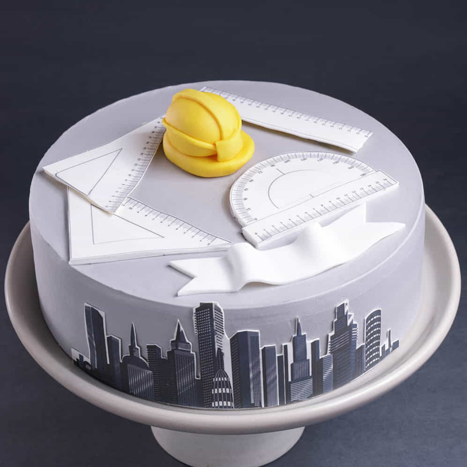 Gray round cake with a city skyline design around the sides, topped with a yellow hard hat and fondant blueprint tools, displayed on a pedestal against a dark background.