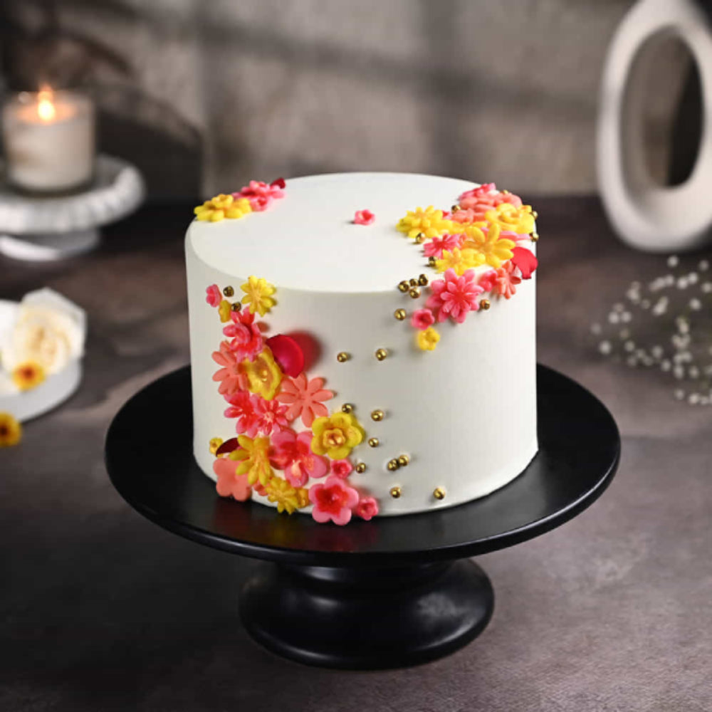 Elegant white designer cake with colorful floral decorations and gold pearls on a black stand, perfect for celebrations in Delhi.