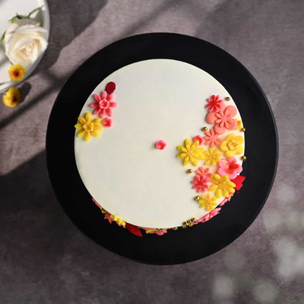 Top view of a white designer cake with pink and yellow floral decorations and gold accents on a black stand, ideal for celebrations in Delhi.