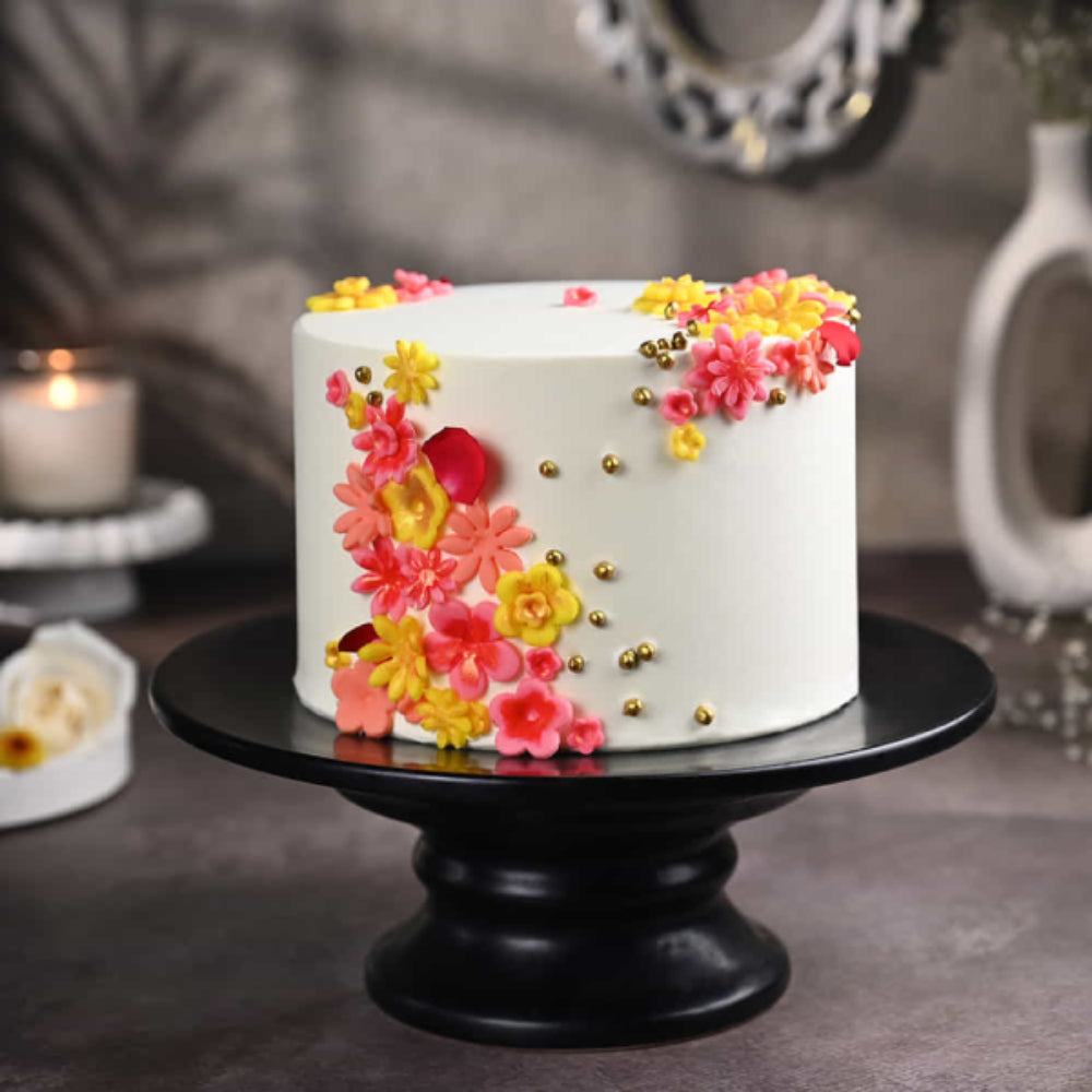 Elegant white designer cake with colorful floral decorations and gold pearls on a black stand, perfect for celebrations in Delhi.
