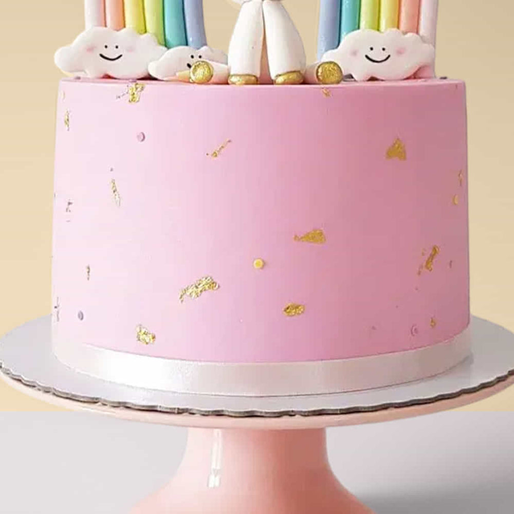 Pink cake with gold decorations and cloud-shaped candles on a stand against a light yellow background