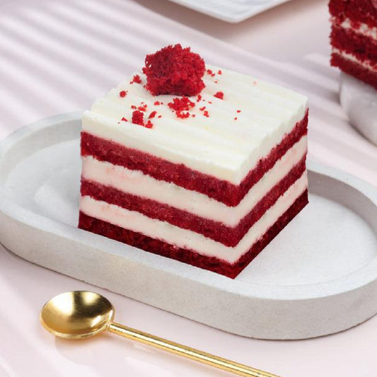 Square red velvet pastry with white cream cheese frosting layers, topped with red crumbs, on a white plate.