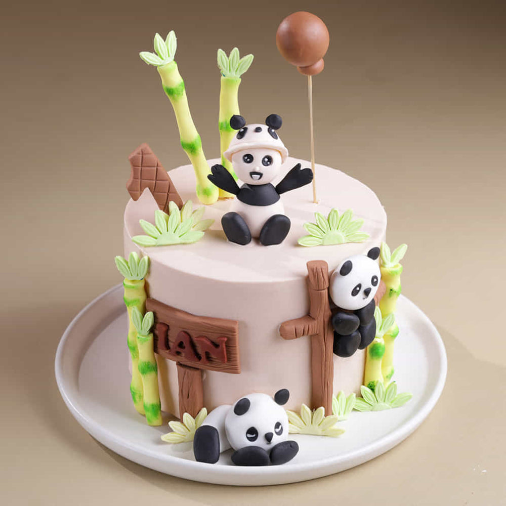 A cream-colored first birthday cake featuring three panda figurines and green bamboo stalks. Top angled view